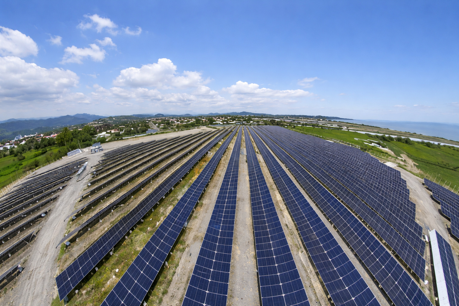Vista aérea de um parque solar com painéis fotovoltaicos alinhados sob céu azul ao meio-dia.