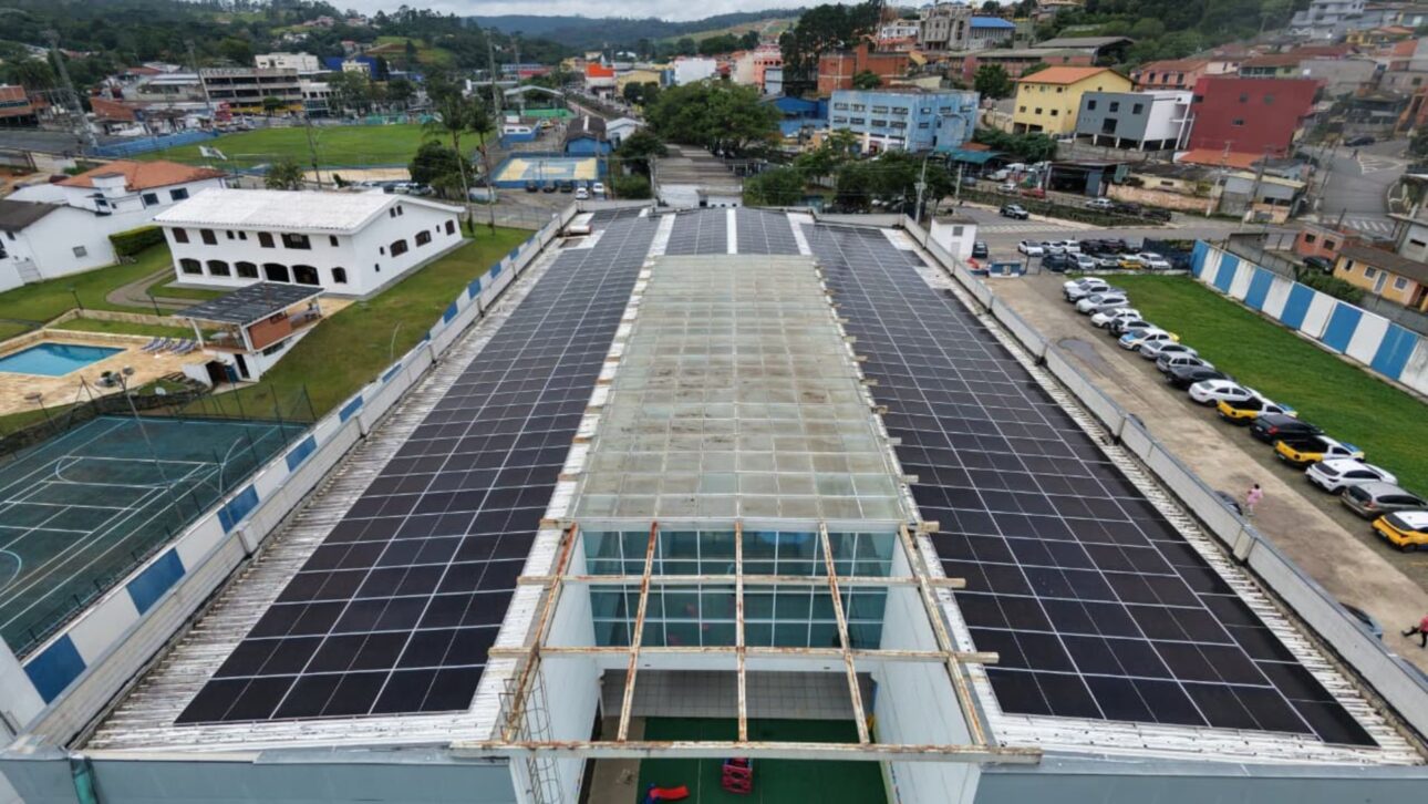 Vista aérea de edificio público con techo cubierto por paneles solares en Cotia, con estacionamiento lateral, cancha deportiva y área urbana alrededor.