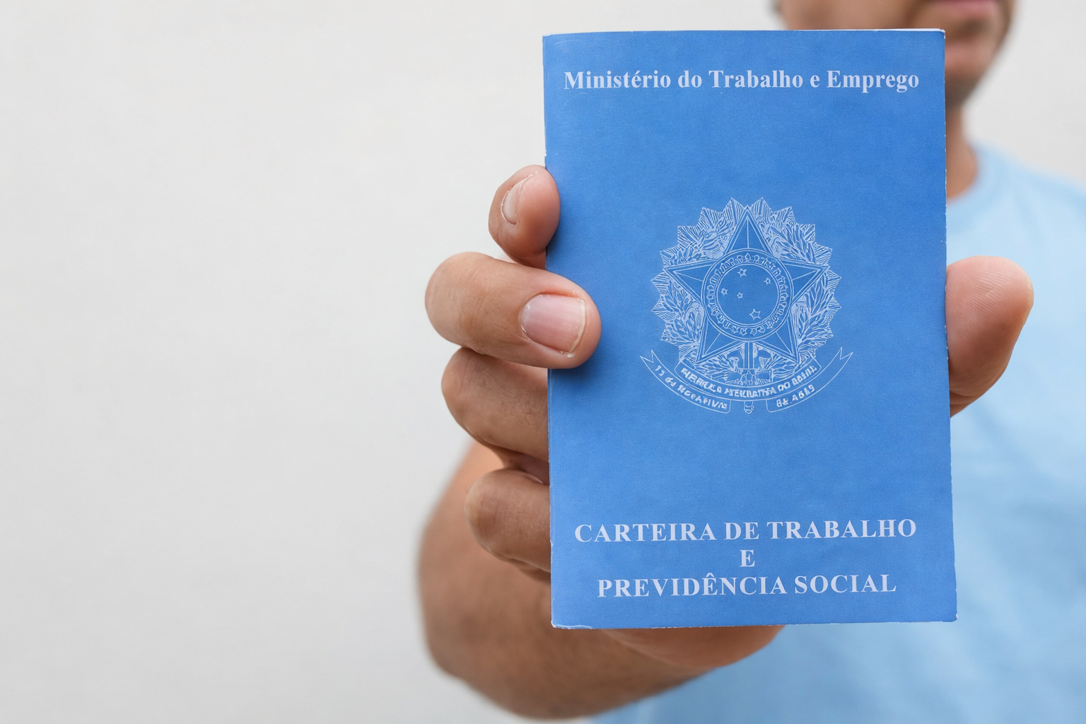 Pessoa segurando uma carteira de trabalho brasileira azul em primeiro plano, usando camisa azul clara, com fundo neutro.