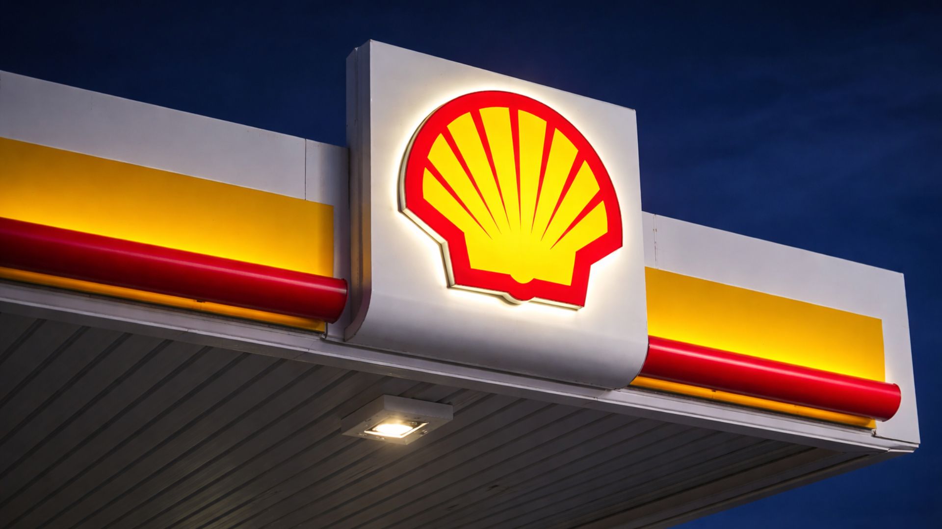 Logotipo iluminado de posto de combustível à noite visto de baixo, com cobertura branca, faixas vermelhas e amarelas e céu azul-escuro ao fundo.