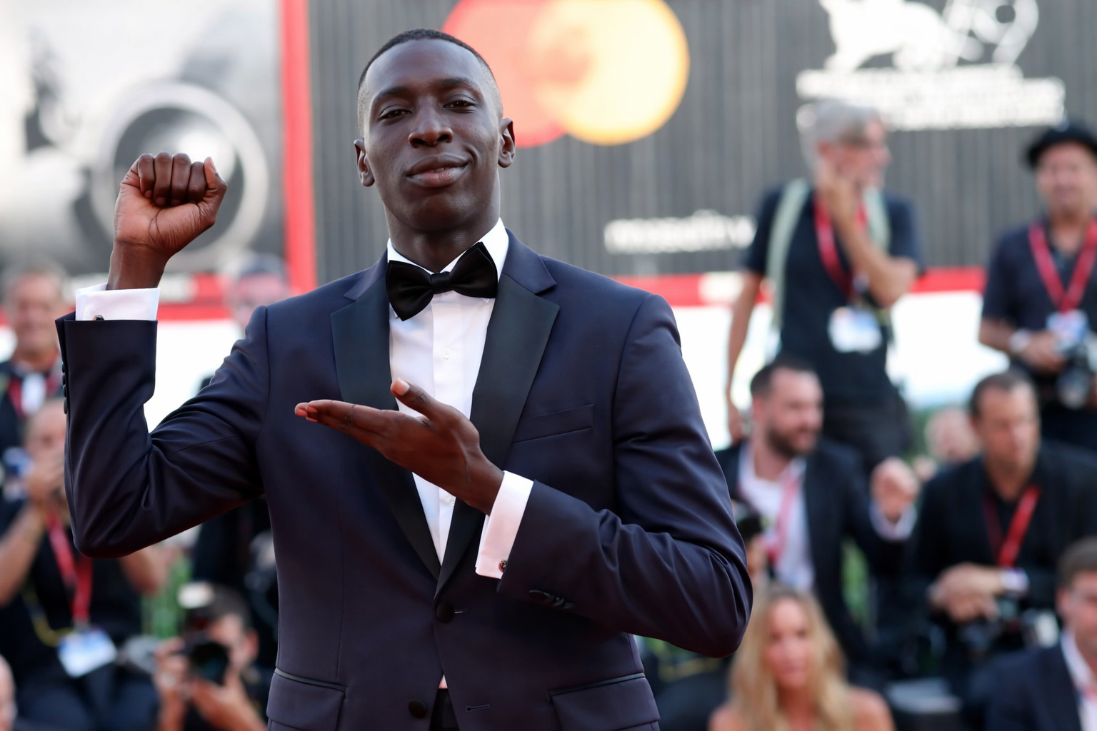 Kaby Lame vestido com terno preto e gravata borboleta faz gesto simbólico no tapete vermelho de um festival internacional de cinema, enquanto fotógrafos registram o momento ao fundo