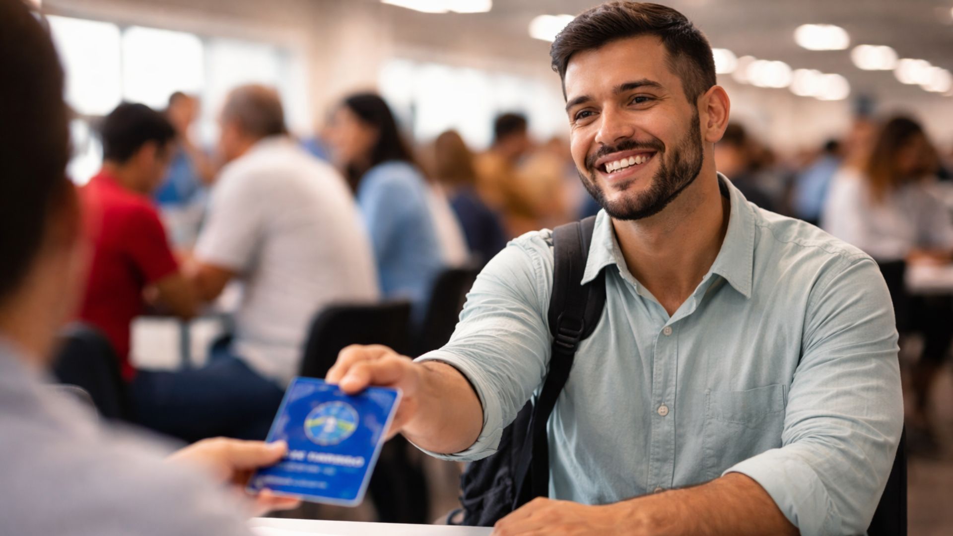 Homem sorrindo entrega carteira de trabalho a recrutador durante mutirão de emprego, com outras pessoas desfocadas ao fundo em atendimento presencial.