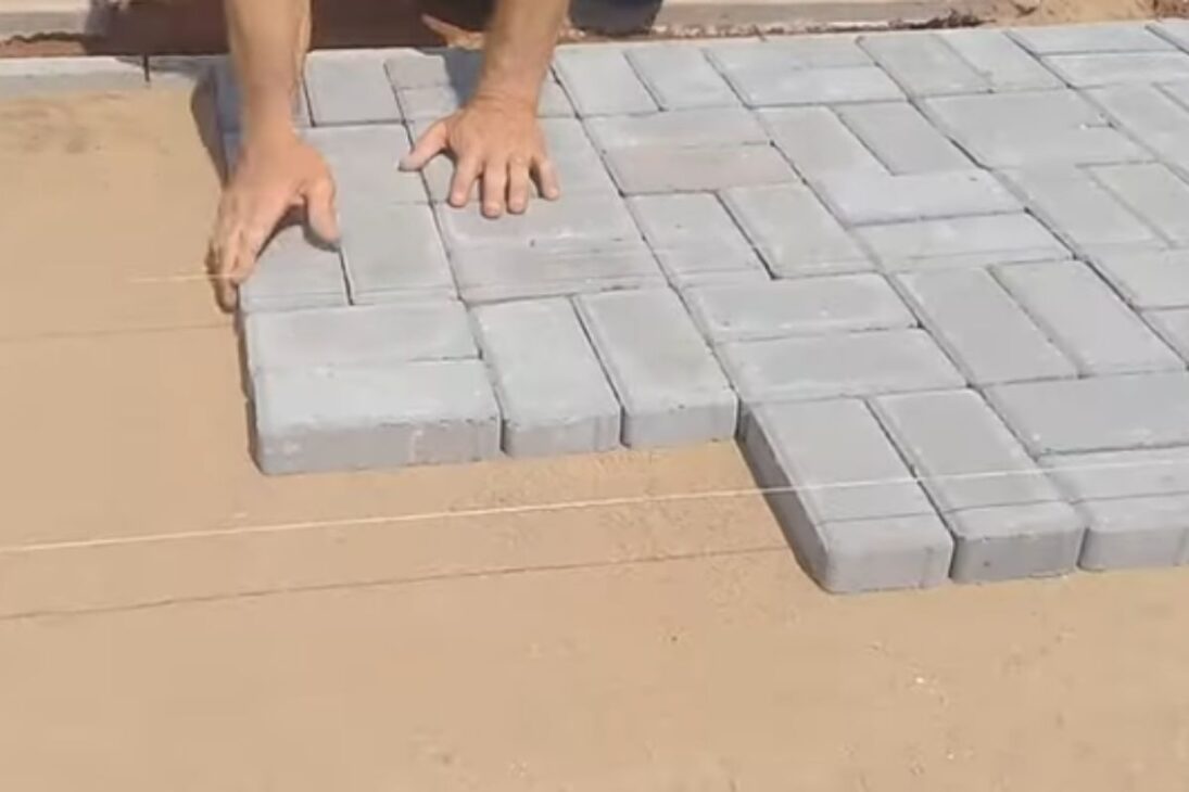 Albañil muestra bloque intertrabado en la acera con arena y placa vibratoria para asegurar juntas y cerrar el acabado.