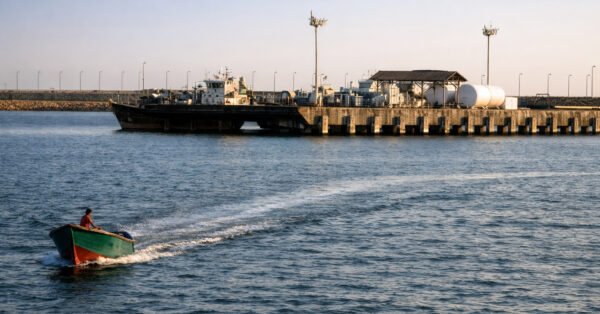 Pequeno barco navegando próximo a um navio petroleiro atracado em terminal marítimo, cenário que representa o transporte de petróleo em rotas estratégicas como o Estreito de Ormuz.