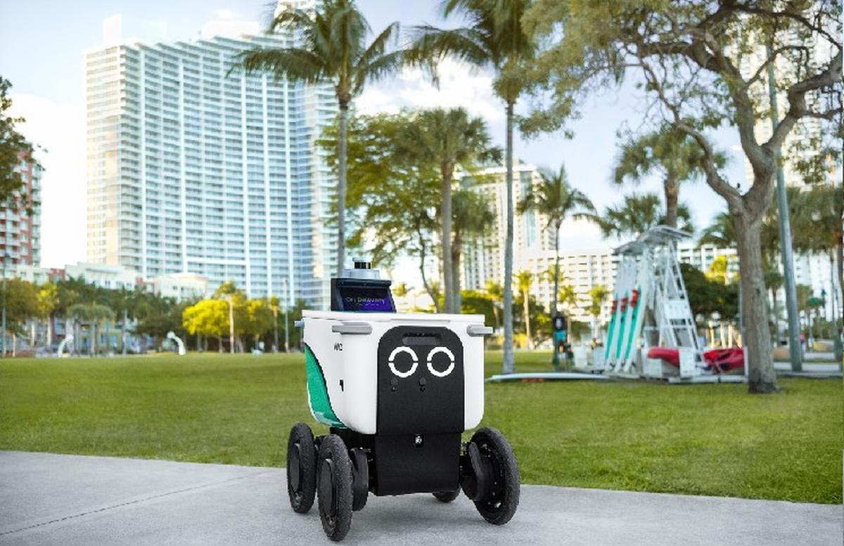 Robô de delivery autônomo circulando em calçada de Miami com prédios e palmeiras ao fundo, representando avanço da tecnologia autônoma na cidade.